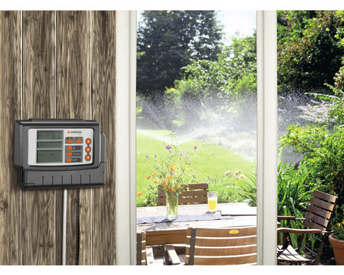Gardena Bewässerungssteuerung zur Gartenbewässerung an einer Holzwand montiert. Im Hintergrund ein Garten mit Rasensprenger und ein Gartentisch.