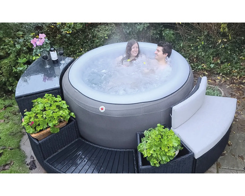 Aufblasbarer Whirlpool im Garten mit zwei Personen, umgeben von modularen Rattanmöbeln mit Tisch, Bank und Pflanzen.