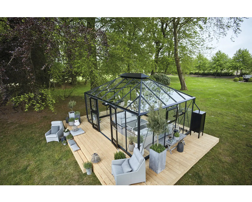 Gewächshaus mit Glaswänden und schwarzem Rahmen auf einer Holzterrasse im Garten, umgeben von Bäumen und Grünflächen.