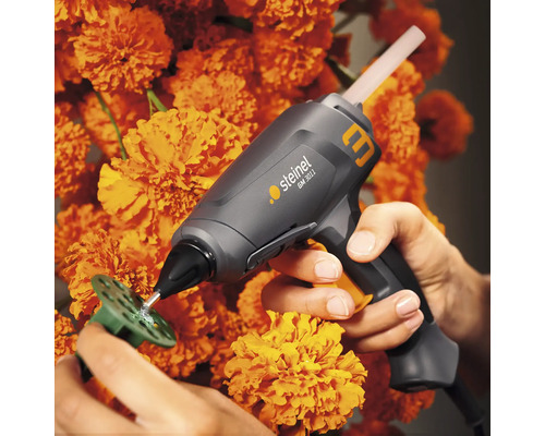 Steinel Heißklebepistole beim Anbringen von Klebstoff auf einer grünen Kunststoffscheibe, umgeben von orangefarbenen Blumen.