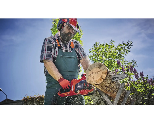 Mann mit Schutzhelm, Gehörschutz und Gesichtsschutz, der im Garten mit einer elektrischen Kettensäge ein Holzstück schneidet.