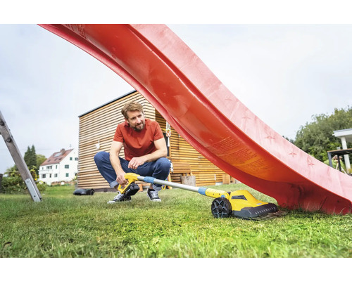 Ein Mann hockt auf einer grünen Wiese und reinigt eine rote Rutsche mit einem gelben Akku-Fugenreiniger.
