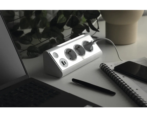 Steckdosenleiste mit drei Schutzkontaktsteckdosen und zwei USB-Anschlüssen auf einem Schreibtisch mit Laptop, Stift, Notizbuch und Smartphone.