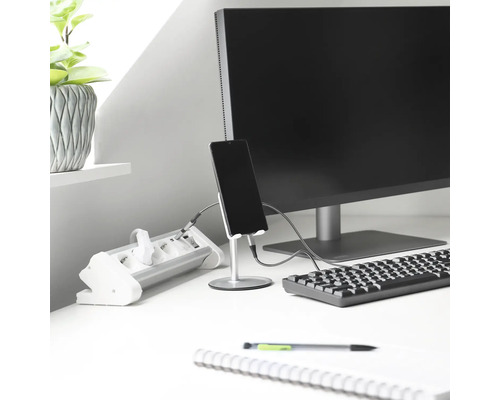 Arbeitsplatz mit Mehrfachsteckdose, Monitor, Tastatur, Stifthalter mit Smartphone, Notizblock und Pflanze