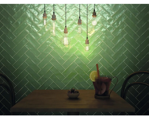 Wandgestaltung mit grünen Fliesen in Fischgrätmuster, beleuchtet mit Glühlampen über einem Tisch mit Getränken