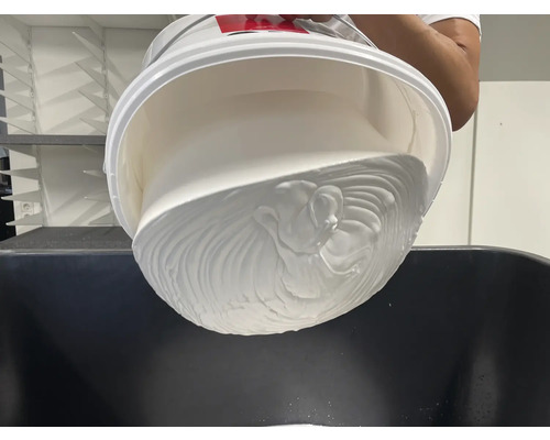 Ein weißer Eimer wird über einen Behälter entleert und gibt eine dicke, weiße Flüssigkeit frei.