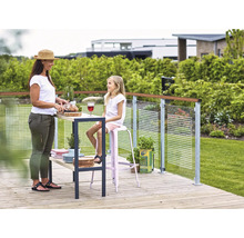 Szene auf einer Holzterrasse mit Mutter und Tochter, die Brot schneidet und auf einem Barhocker sitzt, mit einer Glasbalustrade im Hintergrund